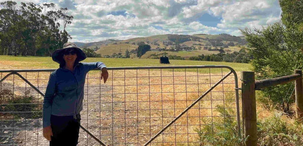 A person wearing a hat and sunglasses leans on a metal gate in front of a grassy field at Marengo Nature Conservation Reserve, with hills and trees visible under a partly cloudy sky in the background.
