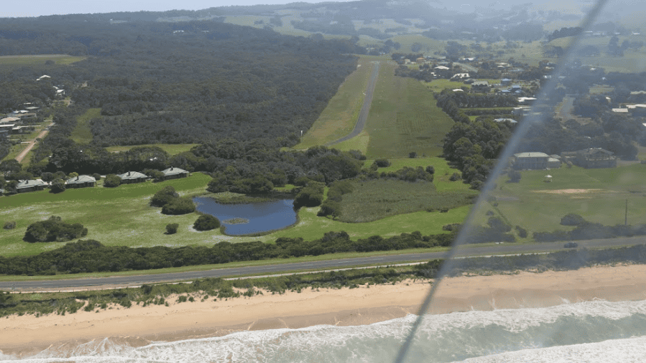 Aerial view of a small airstrip near the coastline, bordered by green fields, a pond, Marengo Nature Conservation Reserve, a beach, and ocean waves, with scattered houses and trees in the background.