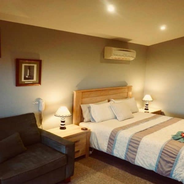 A hotel room with a double bed, neatly folded towels, a gray sofa, bedside tables with lamps, and framed artwork on the walls. The room has gray walls and a ceiling-mounted air conditioner.
