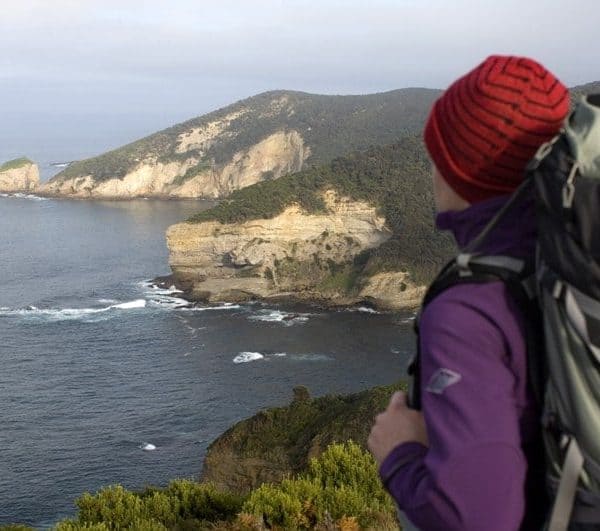 A person with a large backpack and a red beanie looks out at a coastal landscape with cliffs and the ocean.