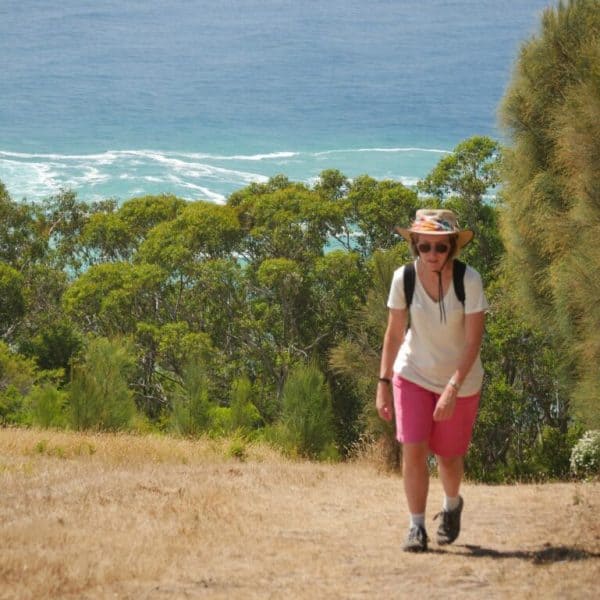 A person wearing a sunhat, sunglasses, a white shirt, and pink shorts hikes uphill on a trail with ocean and trees in the background.