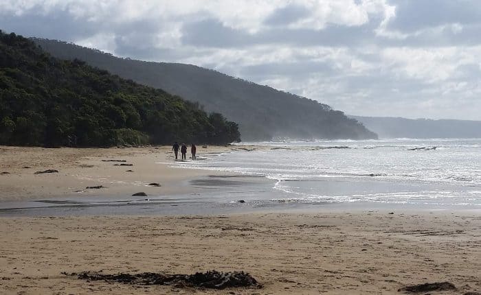Four people walking on a wide, sandy beach with cloudy skies and green hills in the background.