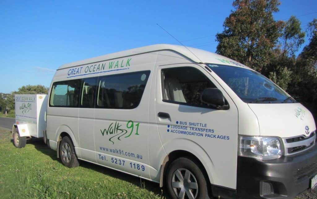 A white van with "Great Ocean Walk" and "Walk 91" branding, parked on a grassy area next to a small trailer. The van offers bus shuttle, luggage transfers, and accommodation packages.