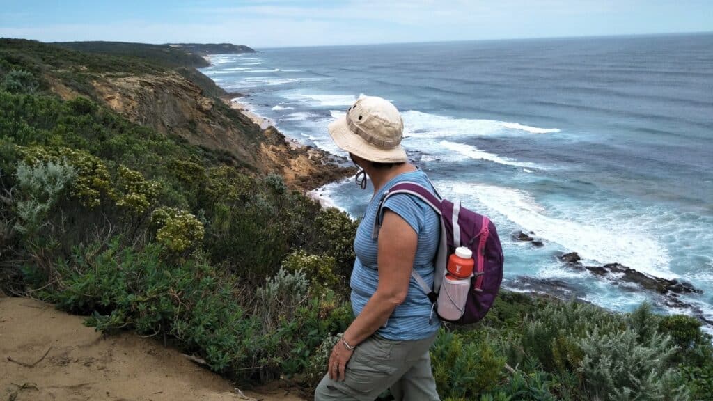 A person wearing a hat, backpack, and hiking gear stands on a coastal trail on the Great Ocean Walk with Walk91 overlooking the ocean and rocky shoreline.