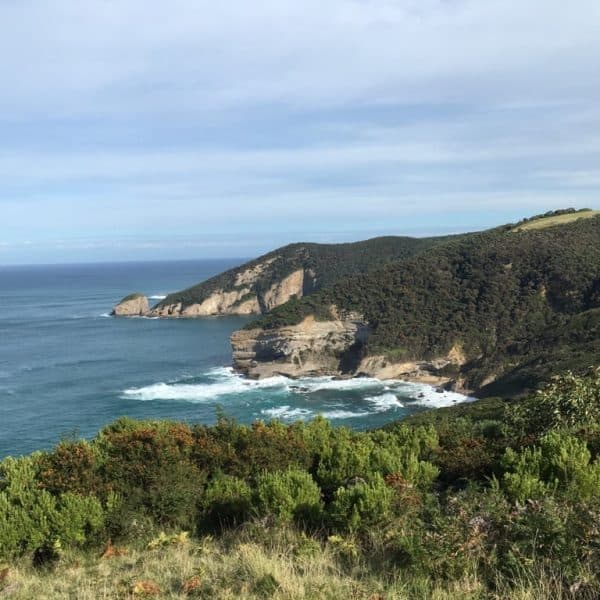 Coastal landscape with cliffs and greenery, overlooking a bay with waves crashing against the shore under a partly cloudy sky.