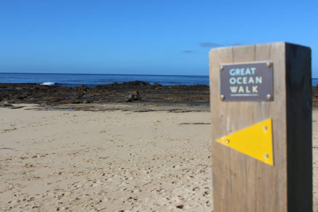 Wooden post with a "Great Ocean Walk" sign and arrow, by a sandy beach with the ocean and rocky shoreline in the background.