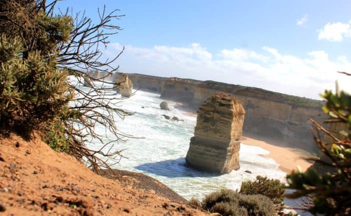 Rock formations stand amid crashing waves along a rugged coastline, with vegetation in the foreground under a clear blue sky.