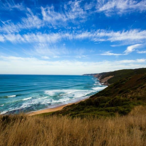 Coastline view featuring blue ocean waves meeting a sandy beach with green hills and grass in the foreground under a sky streaked with wispy clouds.