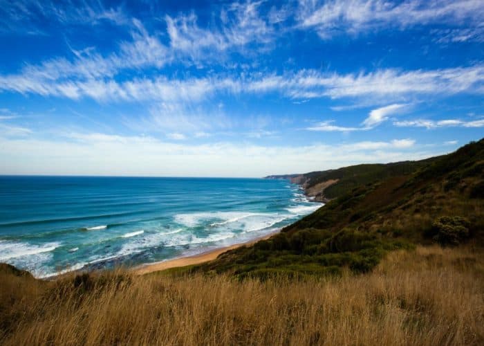Coastline view featuring blue ocean waves meeting a sandy beach with green hills and grass in the foreground under a sky streaked with wispy clouds.