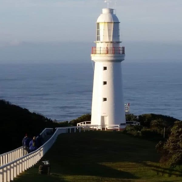 A white lighthouse stands on a grassy hill overlooking the sea. Two people walk along a fenced pathway leading to it.