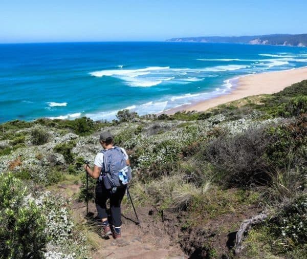 A hiker with a backpack uses trekking poles to walk down a narrow path towards a sandy beach with turquoise waves. Dense greenery surrounds the path, and distant hills extend along the coastline.
