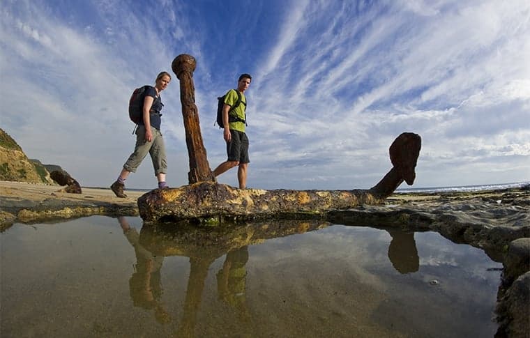 Two hikers walk past a large, rusted anchor partially embedded in the rocky shoreline, Wreck Beach, Great Ocean Walk