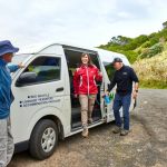 Three people in outdoor clothing are standing around a white shuttle van parked on a rural road. One woman is stepping out of the van while two men stand nearby, one of them assisting her.