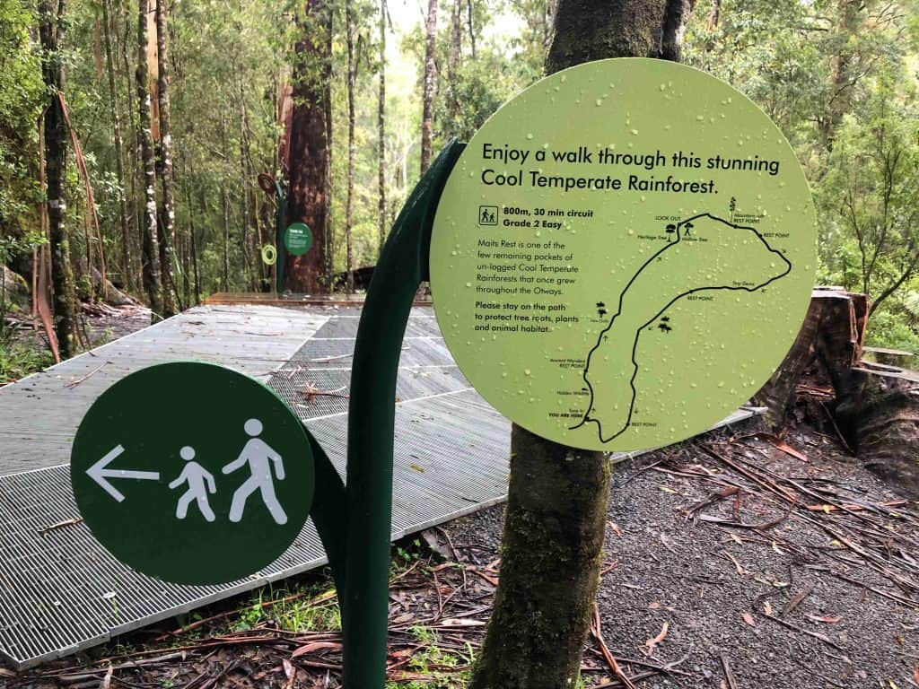 A trail sign in the rainforest points left for walkers, describing a 600m loop through cool temperate forest along the Maits Rest rainforest walk on the Great Ocean Road, with a map and safety information.