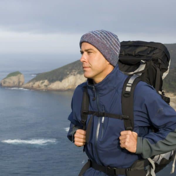 Man wearing a blue jacket, beanie, and backpack, standing on a cliff overlooking the ocean and rugged coastline.