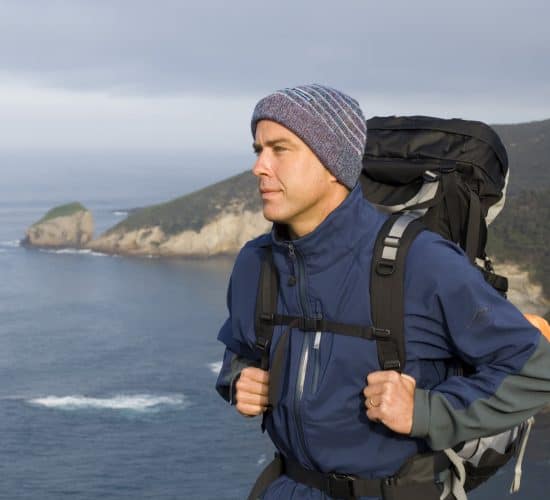 Mark Kininmonth at Walk91 wearing a blue jacket and wool hat stands on a cliff edge overlooking the ocean, with a large backpack on his back. A man wearing a blue jacket and wool hat stands on a cliff edge overlooking the ocean, with a large backpack on his back.