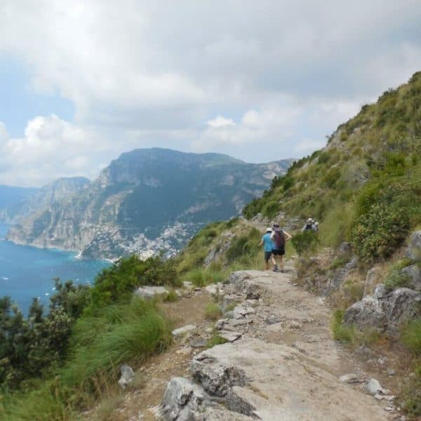A hiker walks along a rocky trail on a hillside overlooking a coastal landscape with cliffs and the sea, under a partly cloudy sky.