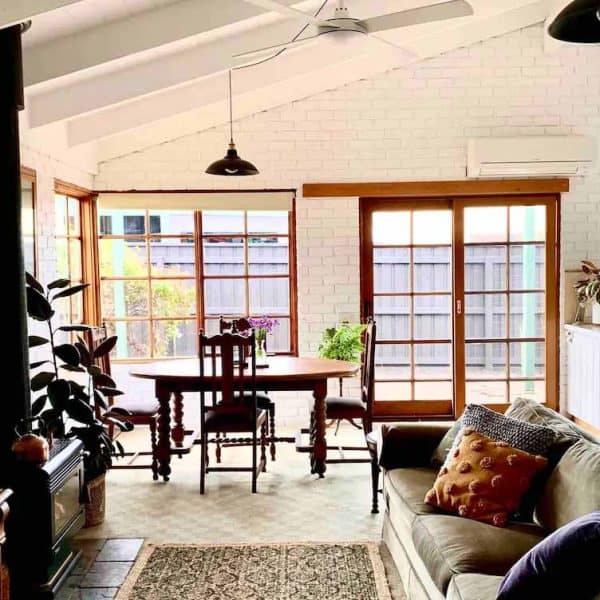 Bright living room with white brick walls, a sofa, a TV, a wood stove, and a dining table. Large windows and glass doors bring in natural light. Open layout includes a kitchen area with white cabinetry.