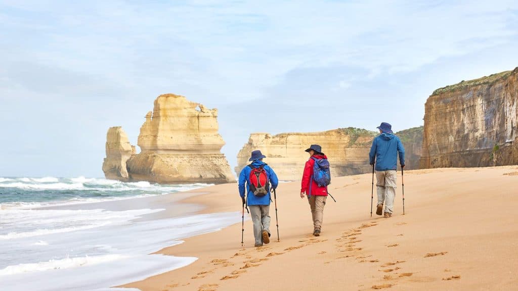 Three walkers from Walk91 walking on the Gibson Steps Beach with the sandstone stack of the 12 Apostles in the distance