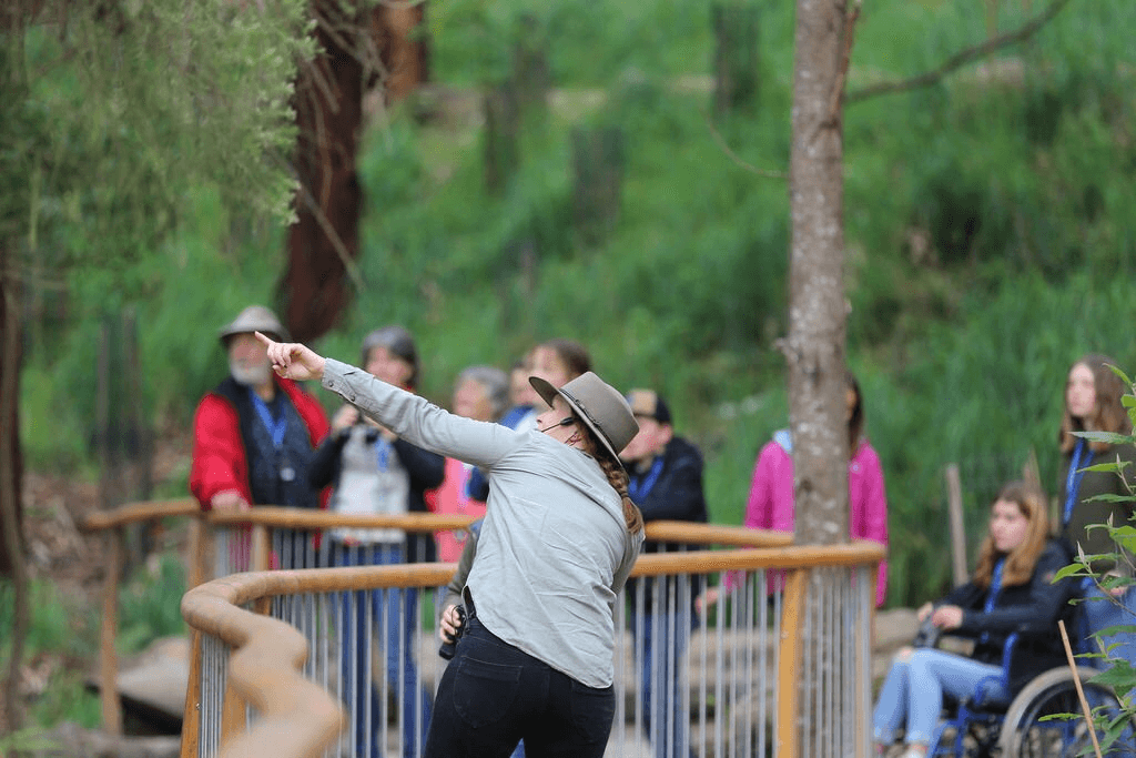 A person in a gray jacket and hat is throwing food over a fence, watched by a group of people including someone in a wheelchair at Wildlife Wonders