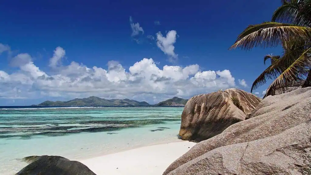 Tropical beach with large rocks on the shore, clear turquoise sea, and distant islands under a partly cloudy sky. Palm trees are visible on the right.