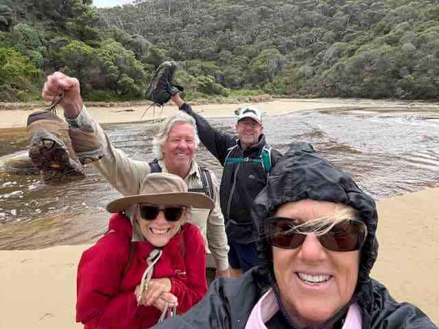 Four adults in outdoor clothing smile for a photo by a riverbank, holding up hiking shoes—celebrating their Self Guided Great Ocean Walk with Walk91—with dense forest in the background.