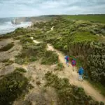 Three hikers walk along a coastal trail surrounded by lush greenery and ocean views under a cloudy sky.