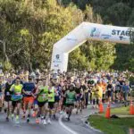 A large group of runners begins a race on the paved Great Ocean Road, passing under a white inflatable start arch, surrounded by trees and spectators—capturing the excitement of the Great Ocean Road Running Festival.