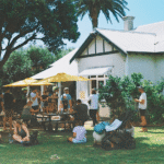 A group of people gather for a wellness event, sitting on the grass and at picnic tables outside a white house with yellow umbrellas on a sunny day along the Great Ocean Road.