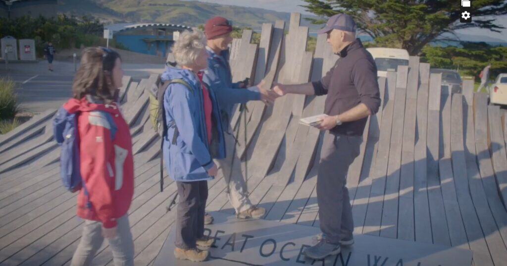 Three people with backpacks meet and shake hands with a fourth on a curved wooden platform outdoors, surrounded by hills and trees, discussing the differences between the Mild Side and the Wild Side of The Great Ocean Walk with Walk91