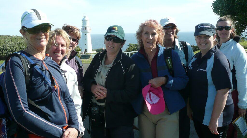 A group of eight women posing outdoors with the white Cape Otway lighthouse and the ocean in the background on a sunny day, whilst enjoying The Great Ocean Walk with Walk91