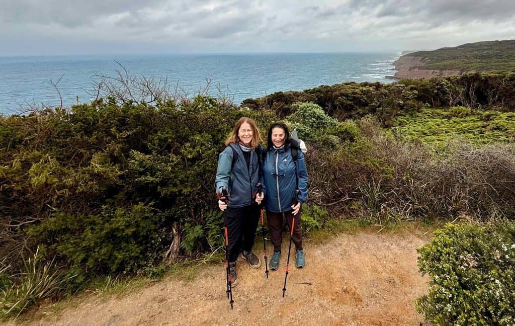 Two people with hiking poles stand on a coastal trail, surrounded by greenery and ocean cliffs beneath a cloudy sky—capturing the spirit of adventure in Walk91’s 10 Essential Tips for Hiking the Great Ocean Walk.