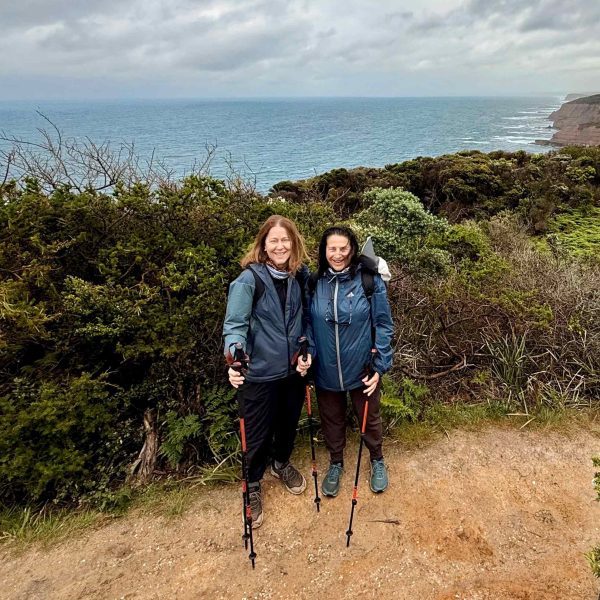 Two people with hiking poles stand on a coastal trail, surrounded by greenery and ocean cliffs beneath a cloudy sky—capturing the spirit of adventure in Walk91’s 10 Essential Tips for Hiking the Great Ocean Walk.