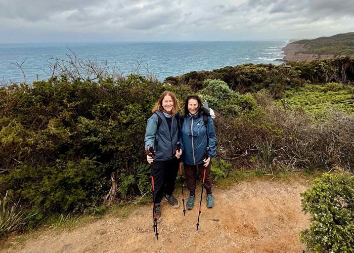 Two people with hiking poles stand on a coastal trail, surrounded by greenery and ocean cliffs beneath a cloudy sky—capturing the spirit of adventure in Walk91’s 10 Essential Tips for Hiking the Great Ocean Walk.