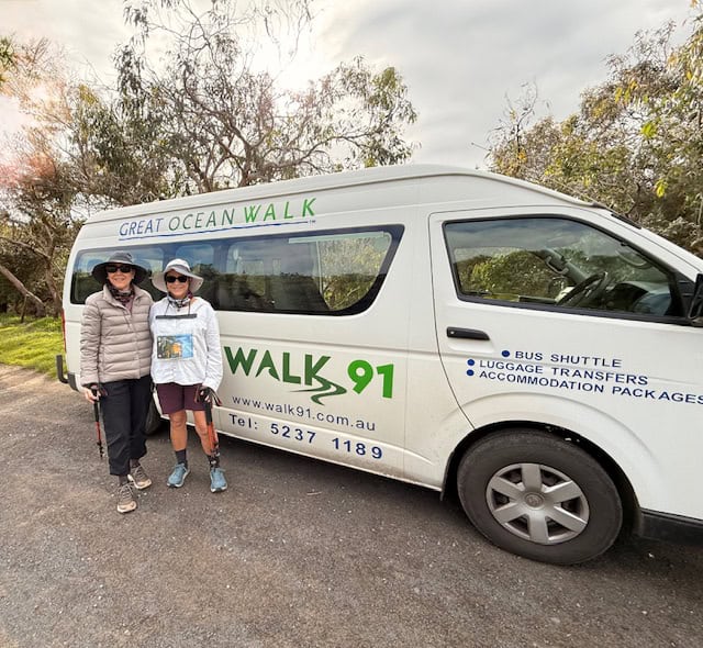 Two people wearing hats and sunglasses stand beside a white Walk91 van with Great Ocean Walk hike signage, parked on a paved road near trees.