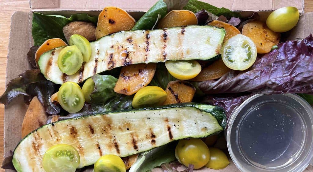A takeout container with grilled zucchini slices, yellow cherry tomatoes, roasted yellow beets, mixed greens, and a small cup of vinaigrette