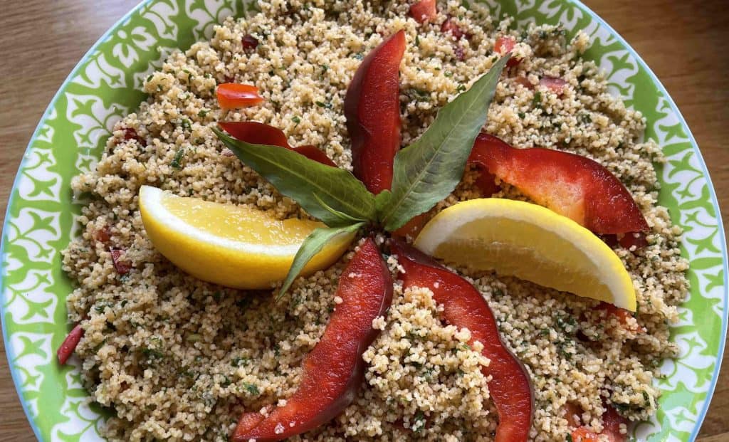 A plate of couscous salad garnished with red bell pepper slices, lemon wedges, and fresh basil leaves on a green and white patterned dish