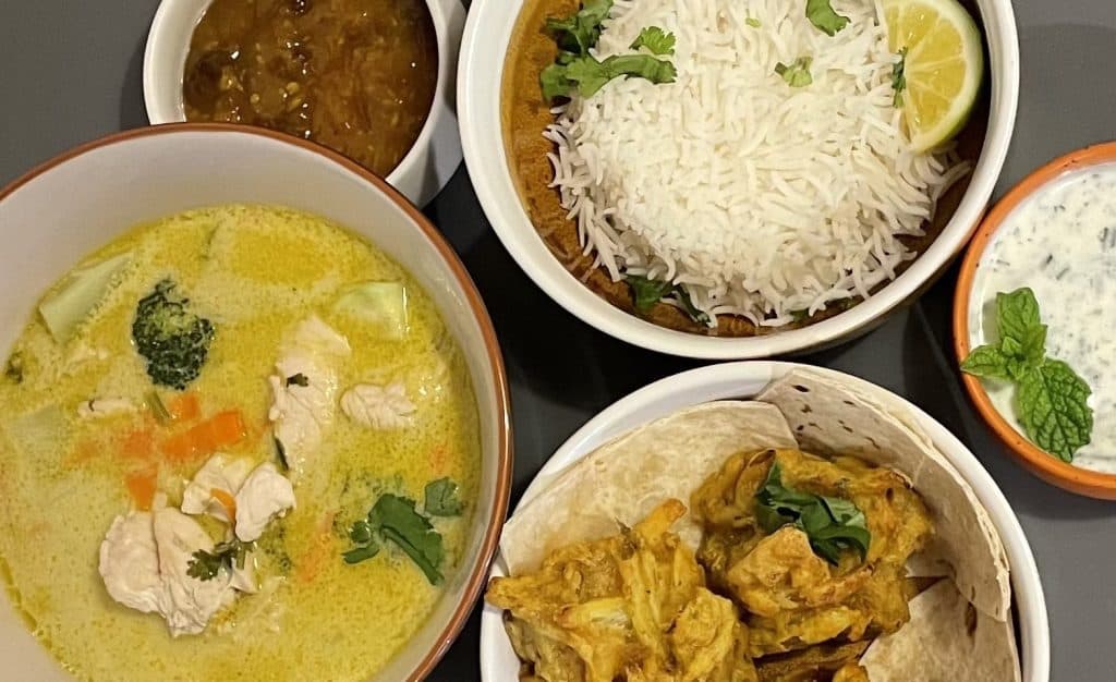 A meal featuring white rice with a lemon wedge, chicken curry, chicken in yellow sauce with vegetables, naan, chutney, and raita