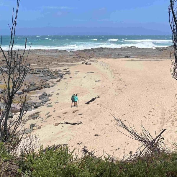 Two people walk along a sandy beach bordered by rocks and sparse trees, with waves crashing in the distance under a partly cloudy sky—much like scenes found along the Great Ocean Walk. Which Great Ocean Walk Map Should You Use? Ask Walk91 for tips!.