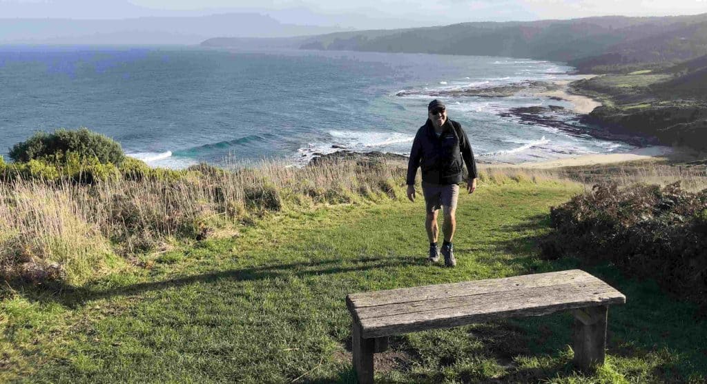 A man wearing a jacket and shorts walks uphill on a grassy path near a wooden bench, with the ocean and coastline in the background—just one of many scenic spots you'll find on the Great Ocean Walk. Which Great Ocean Walk Map Should You Use? | Walk91.