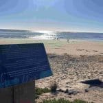 An information sign at the beach overlooks the sea, whilst a few people walk along the sandy shore under a bright, sunny sky, marking the easiest section of the Great Ocean Walk from Apollo Bay to Shelly Beach.