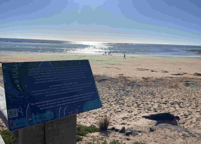An information sign at the beach overlooks the sea, whilst a few people walk along the sandy shore under a bright, sunny sky, marking the easiest section of the Great Ocean Walk from Apollo Bay to Shelly Beach.